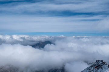 A breathtaking winter mountain landscape covered in deep snow, with dramatic clouds rolling over rugged alpine peaks under a bright blue sky, creating a serene and powerful natural atmosphere.