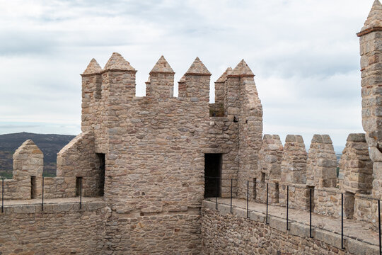 Detalhe da arquitetura militar do castelo de Penedono com foco nas ameias piramidais e no caminho de ronda em alvenaria de pedra sob um c&eacute;u nublado, destacando o topo da muralha medieval em Portugal