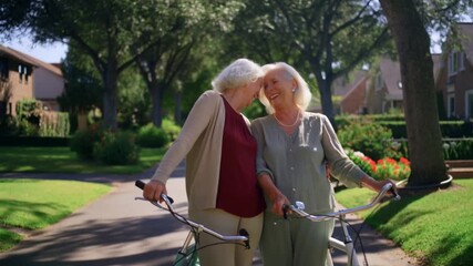 Two senior women sharing an intimate moment while walking their bicycles down a path. The scene is set in a peaceful neighborhood. Stock Video