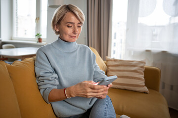 Positive middle aged woman resting on couch in living room with phone, scrolling social networks...