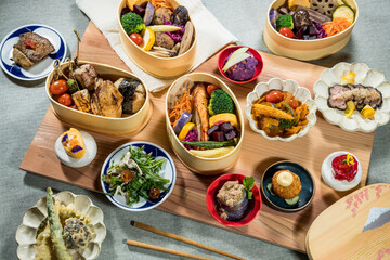 A Japanese-style meal with bento boxes and side dishes including grilled fish, rice balls, vegetables, tempura, salad, and desserts.