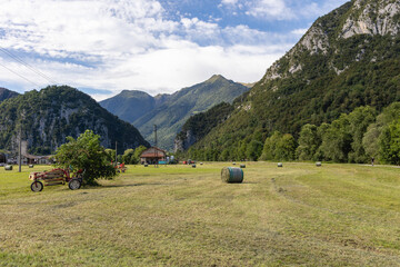 Rural alpine landscape in Valsassina, Lombardy, Italy. Idyllic view of the valley with agricultural fields, hay bales and equipment, surrounded by lush green mountains under a cloudy sky.