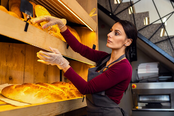 Smiling female baker arranging freshly baked bread on shelves inside bakery.