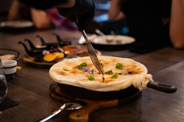 A fine dining dish being served in a modern restaurant — a baked pan covered with golden crust, garnished with edible flowers and herbs, as a chef slices through the top to reveal the steaming aroma.
