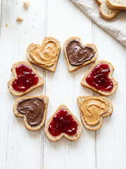 Heart-shaped bread slices with peanut butter, chocolate, and jam arranged in a circle on white wood. A creative food concept for love, breakfast, or sweet homemade moments.