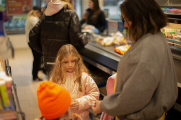 Young Girl Paying At Checkout Register, White Mother Nearby Guiding Purchase While Child Handles Item Bright Snack Displays, Orange Beanie On Nearby