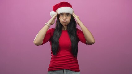 Young hispanic woman in red top holds santa hat with both hands, puffs cheeks and looks annoyed in studio; holiday frustration.