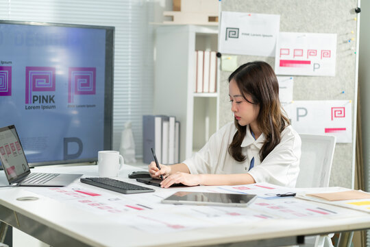 Asian female graphic designer or brand consultant working on a logo design using a digital pen tablet and computer monitor in modern studio office Concept of creative workflow and professional design - Powered by Adobe
