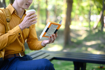 Cropped shot man sits on a bench in a green park reading a book with a coffee cup