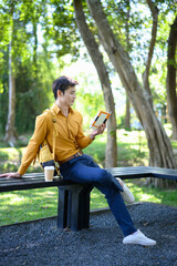 A man sits on a bench in a green park reading a book with a coffee cup