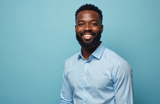 African american man with beard smiles warmly. Young adult male wears light blue collared shirt against solid blue background. Confident person ready for business success. Looks forward with positive