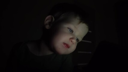 a little boy in the dark watches cartoons on a tablet, his face softly lit by the screens glow, capturing a quiet, cozy moment of nighttime entertainment and digital immersion