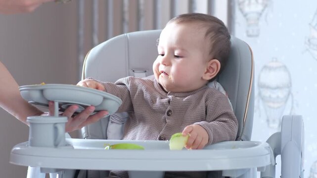 Baby Eating From Highchair, Mother Feeding, Baby Explores Avocado Puree With Hands While Caregiver Offers Spoon, Messy Playful Moments Under Soft