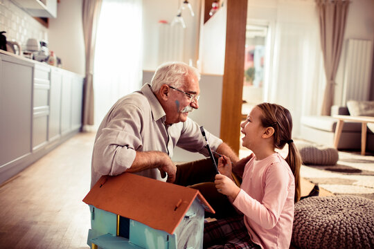 Senior grandfather and child granddaughter painting a dollhouse and laughing at home