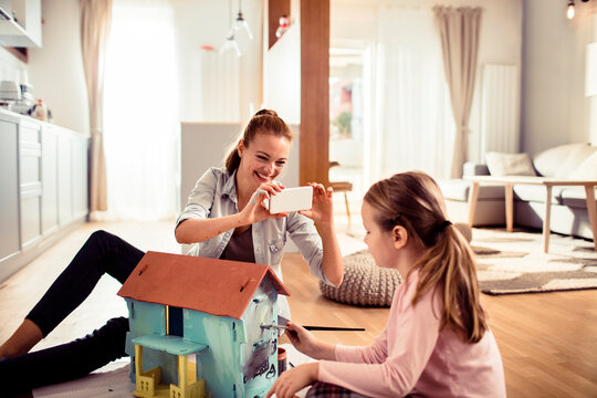 Adult mother photographing young daughter painting dollhouse in living room, happy