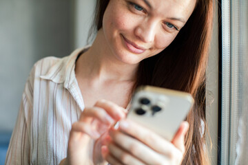 Adult woman texting on smartphone by window at home, relaxed smile