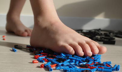 Obraz premium A child's bare foot steps on the scattered parts of a construction kit on the floor. Close-up, parquet, natural light, home scene, emphasis on safety.
