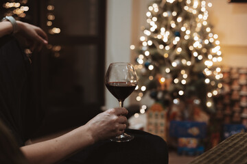 Cozy Christmas evening scene, a person holds a glass of rich red wine against a background of a decorated festive tree with sparkling bokeh lights.