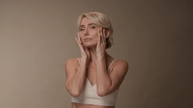 Static shot of blonde mature woman doing daily facial massage while admiring herself