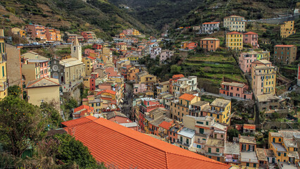 The colorful houses of Riomaggiore, a cozy town in Cinque Terre, descend to a narrow street, surrounded by green terraces and mountains. The atmosphere is warm, colorful, and Mediterranean.