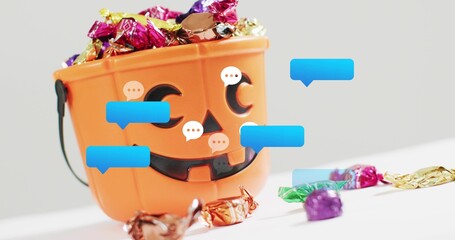 Fototapeta premium Sitting jack-o'-lantern bucket holding foil candies and wrappers on white table with chat bubbles