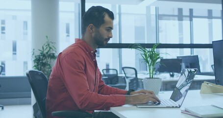 Typing Middle Eastern man in red shirt and dark pants at white desk using silver laptop © vectorfusionart