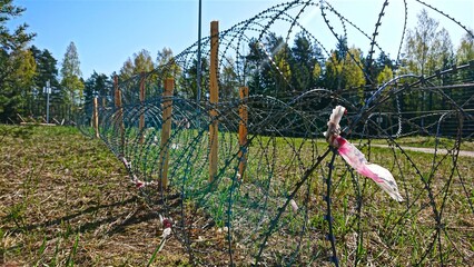 A long razor wire fence stretches across a grassy field near a lush woodland area under a clear...