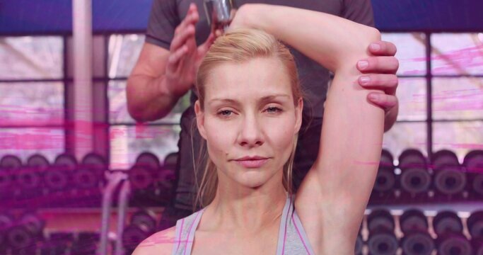 Female client receiving trainer guiding arm behind head in gym, light gray tank top, dumbbell rack - Powered by Adobe