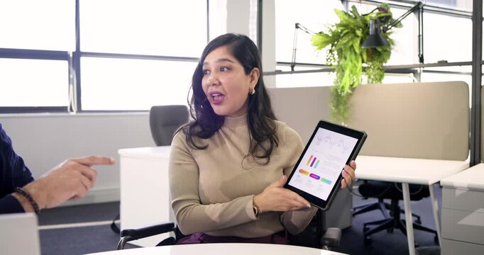 Mid-adult Hispanic woman at meeting table lifting tablet showing charts then presenting report