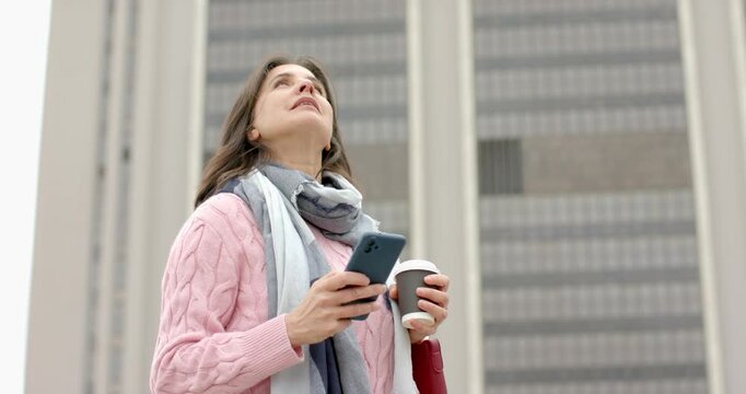 Mature Hispanic woman gazing upward scanning facade checking phone sipping coffee outside tower