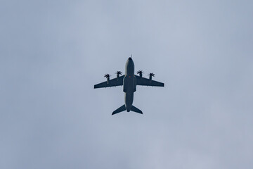 Four-engine turboprop transport aircraft seen from below against a pale sky. Clean silhouette with wide wings and spinning propellers overhead.
