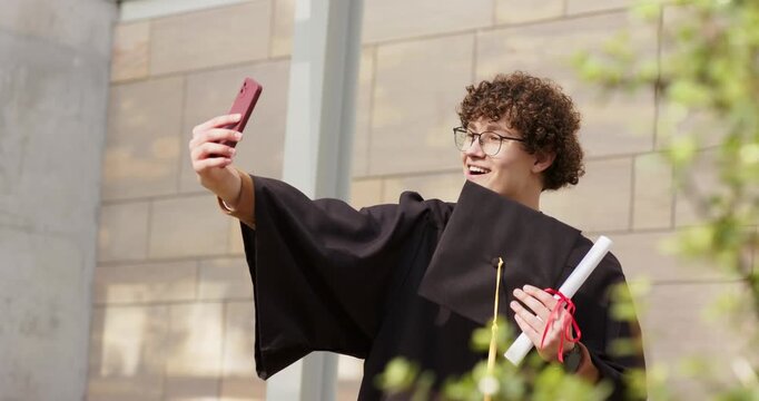 After graduation male grad student holding mortarboard and diploma taking phone selfie on campus