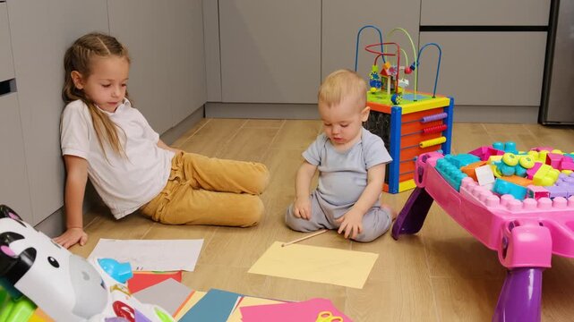 The older sister supervise the younger brother playing together Happy childhood, charming little kids drawing by colored pencils on floor in cozy living room, 