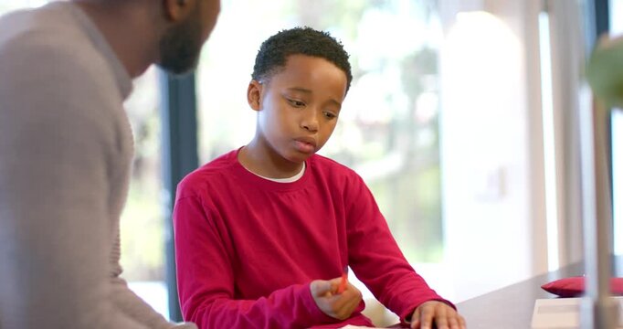 African American father guiding son adjusting pencil, writing homework in notebook at dining table