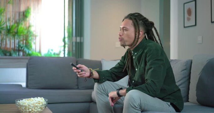 Mid adult man pressing remote seeking show, grabbing popcorn and surfing channels in living room