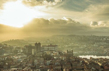 Rooftops gleam as a sunbeam strikes buildings across Vila Nova de Gaia in Portugal. Hazy morning light softens the landscape beneath a sky brushed with ethereal clouds