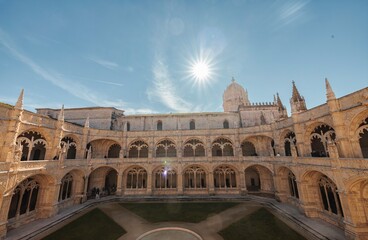 Fototapeta premium Sunlight streams into the courtyard of the Jeronimos Monastery in Lisbon. The ornate architecture of the cloisters gleams under the bright, clear sky during the daytime