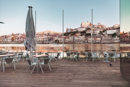On a wooden deck, empty tables and chairs stand still. Douro river softly reflects morning light with Vila Nova de Gaia's mirrored building gleaming, Portugal