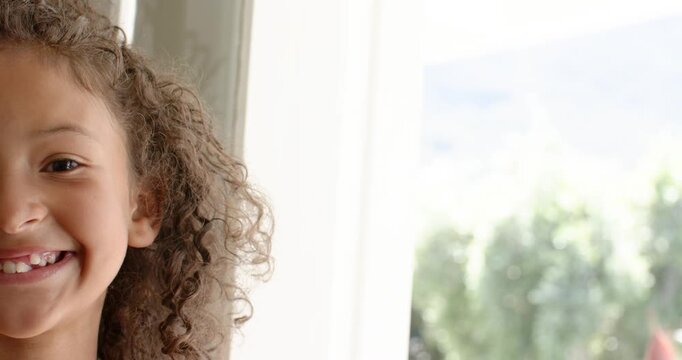 Schoolgirl noticing camera smiling and blinking beside curtain-covered window at home, copy space