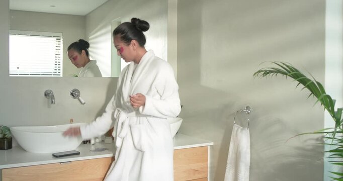 Asian woman at sink reaching eye patch jar, applying patches, scrolling phone, smiling copy space