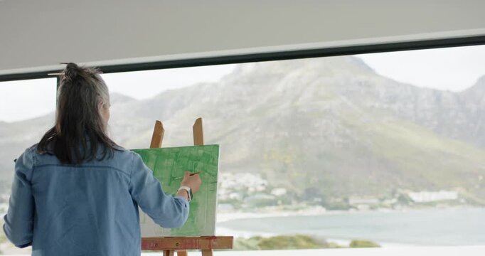 Senior female artist is dipping brush in green paint, painting canvas, pausing to survey bay view