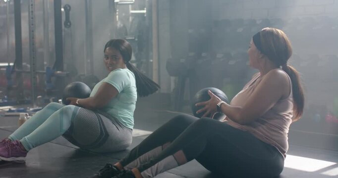 Diverse female friends initiating Russian twists on mats in gym with med balls for core workout