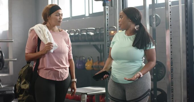 Diverse female friends entering gym area carrying bag and towel, sharing smartphone workout tips