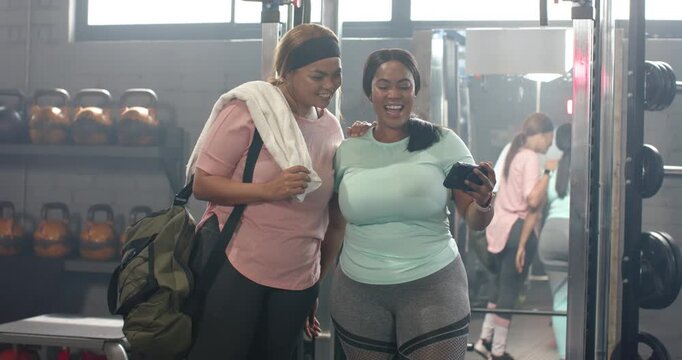 Mid adult female friends leaning by gym mirror, reacting to phone content, holding duffel and towel