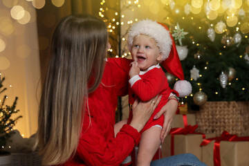 Mother with her baby in pajamas and Santa hat near Christmas tree