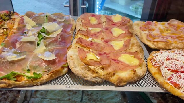 assorted artisan pizzas on glass counter, rustic pizzeria display with melted mozzarella, prosciutto slices, pineapple chunks,