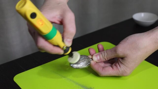 Man using a hand sander with felt disc to clean plaque from a metal spoon on a green mat. Polishing silverware restoration process.
