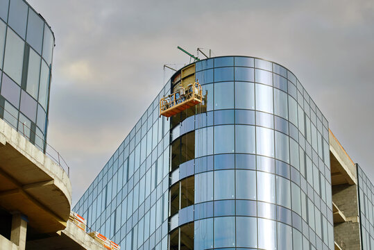 Glass building under construction, sweeping curved facade, incomplete glass walls under moody overcast sky with workers on suspended platforms. Worker on suspended cradle installing cladding panels