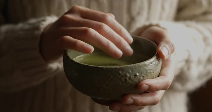Hands holding warm matcha tea bowl wearing knitted sweater.