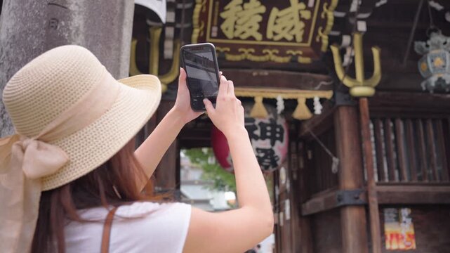 Slow motion of a woman with a sunhat photographing a Japanese temple with her phone. She stands in front of wooden structures capturing architectural details.